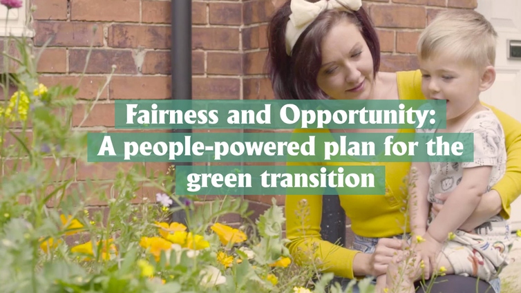 A fair skinned woman with dark red hair, wearing a yellow top, is holding a small boy with blond hair, next to a bunch of daffodils and green plants.