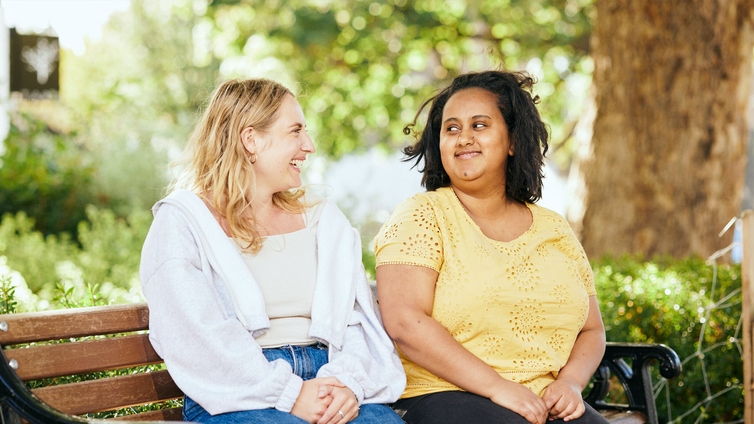 Two women sitting on a wooden bench, smiling at each other. The person on the left has blond hair and is wearing a white jacket. The person on the right has black hair and is wearing a yellow top. A tree can be seen in the background.